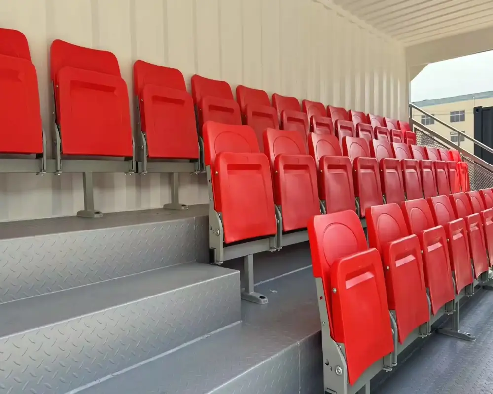 Seating: Several rows of bright red, fold-up plastic seats are mounted on a grey tiered structure. Structure: The seats are installed on light grey metal or concrete risers. The background consists of a beige or off-white corrugated wall, suggesting a modular or temporary grandstand design. Layout: The rows are arranged in a tiered, "bleacher" style to provide clear sightlines. Indicator: In the top right corner, there is a blue square icon with a white checkmark, often used to signify that a selection has been made or a task is complete.