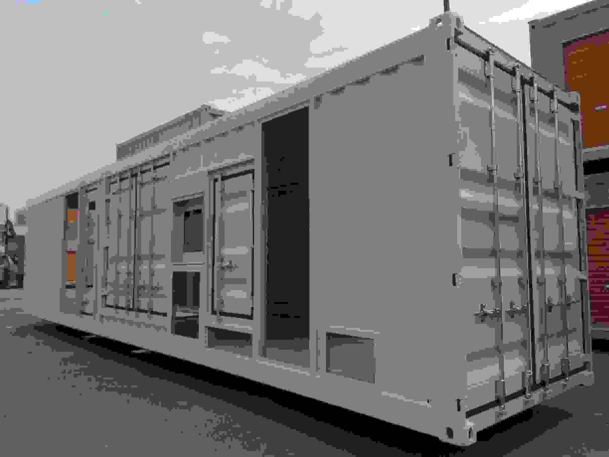 Teal green genset container with white frame borders, single hinged door with latches, elevated on orange adjustable leveling feet, standing in an industrial storage yard alongside similar units, providing secure housing for diesel generator sets