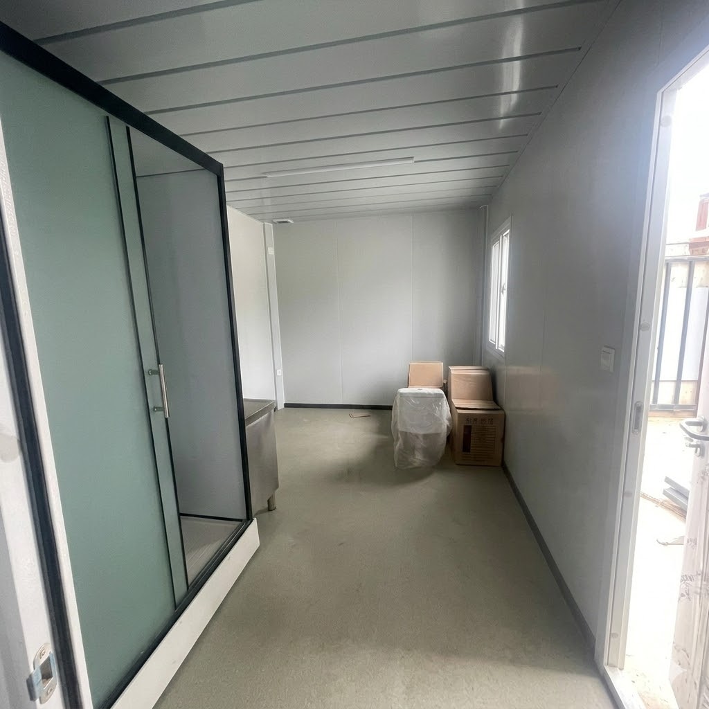 Interior view of a modern prefabricated container house featuring a glass shower cabin, compact living space, and gray panel walls.