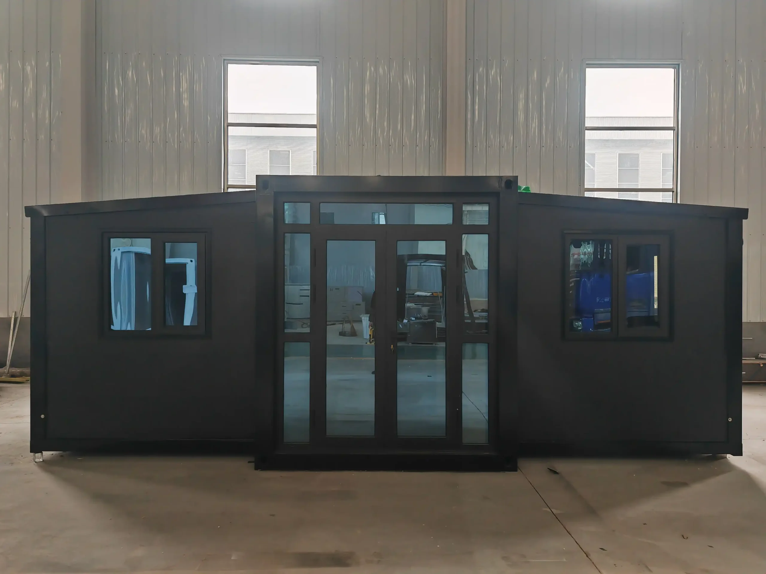Vista externa de uma casa modular expansível em contêiner preta e moderna instalada em um cenário de galpão industrial, apresentando grandes portas duplas de vidro e janelas isoladas.