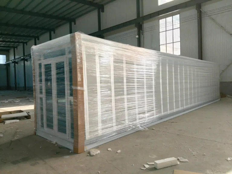 A completed white Malaysia expandable container house project featuring a large integrated front porch with white railings and stairs, set on an artificial grass surface within a professional assembly facility.