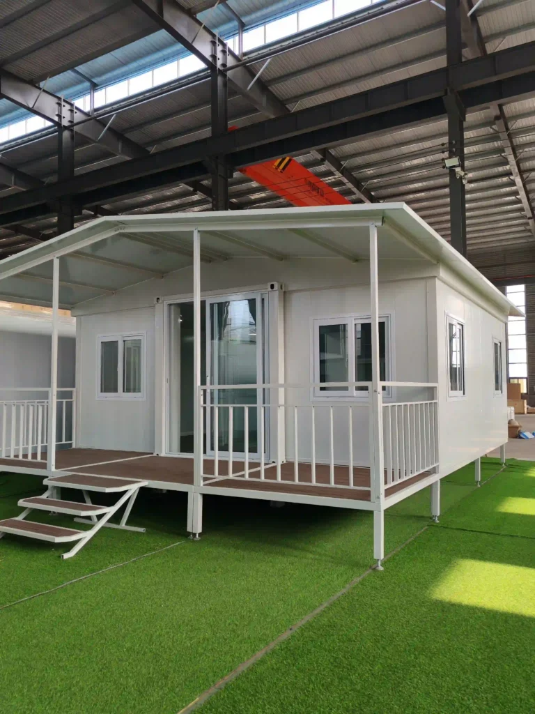 A completed white Malaysia expandable container house project featuring a large integrated front porch with white railings and stairs, set on an artificial grass surface within a professional assembly facility.