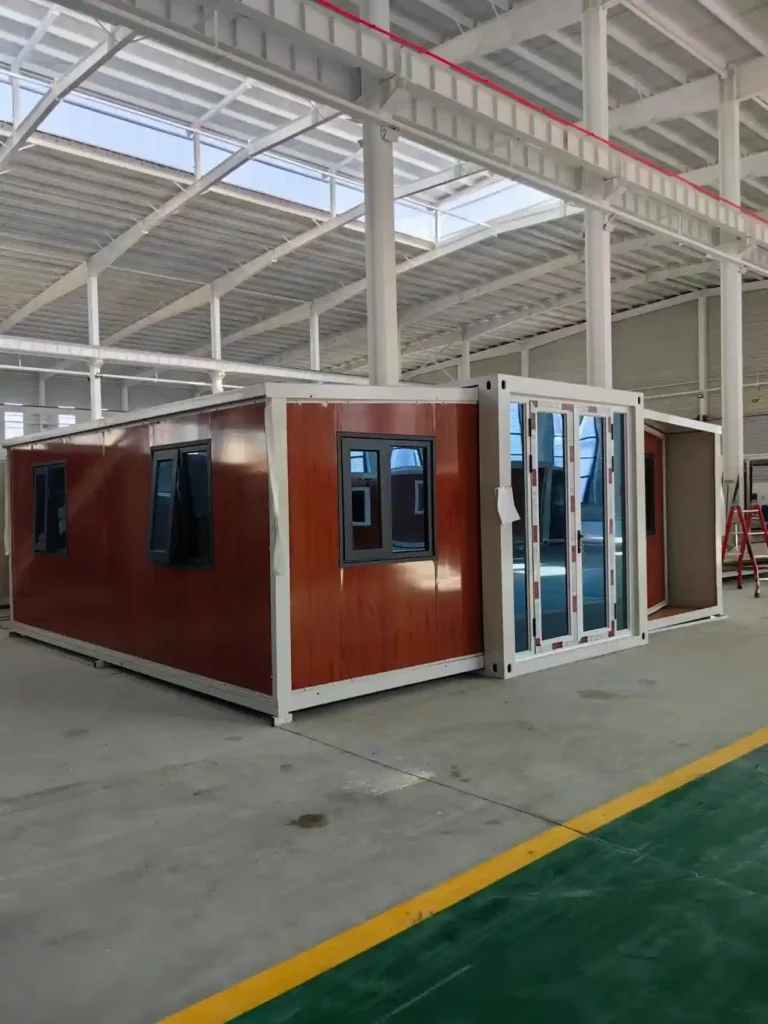 A brown wood-grain finish expandable container house with glass sliding doors and black-framed windows, displayed inside a manufacturing warehouse for a project in Poland.