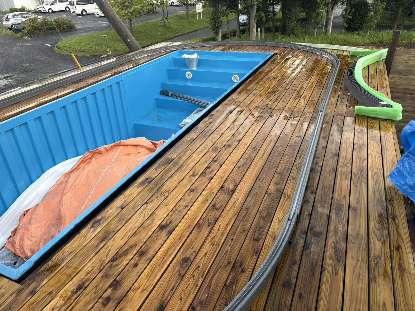 A bright blue container pool under construction, integrated into a polished wooden deck with a curved track system for a sliding cover.