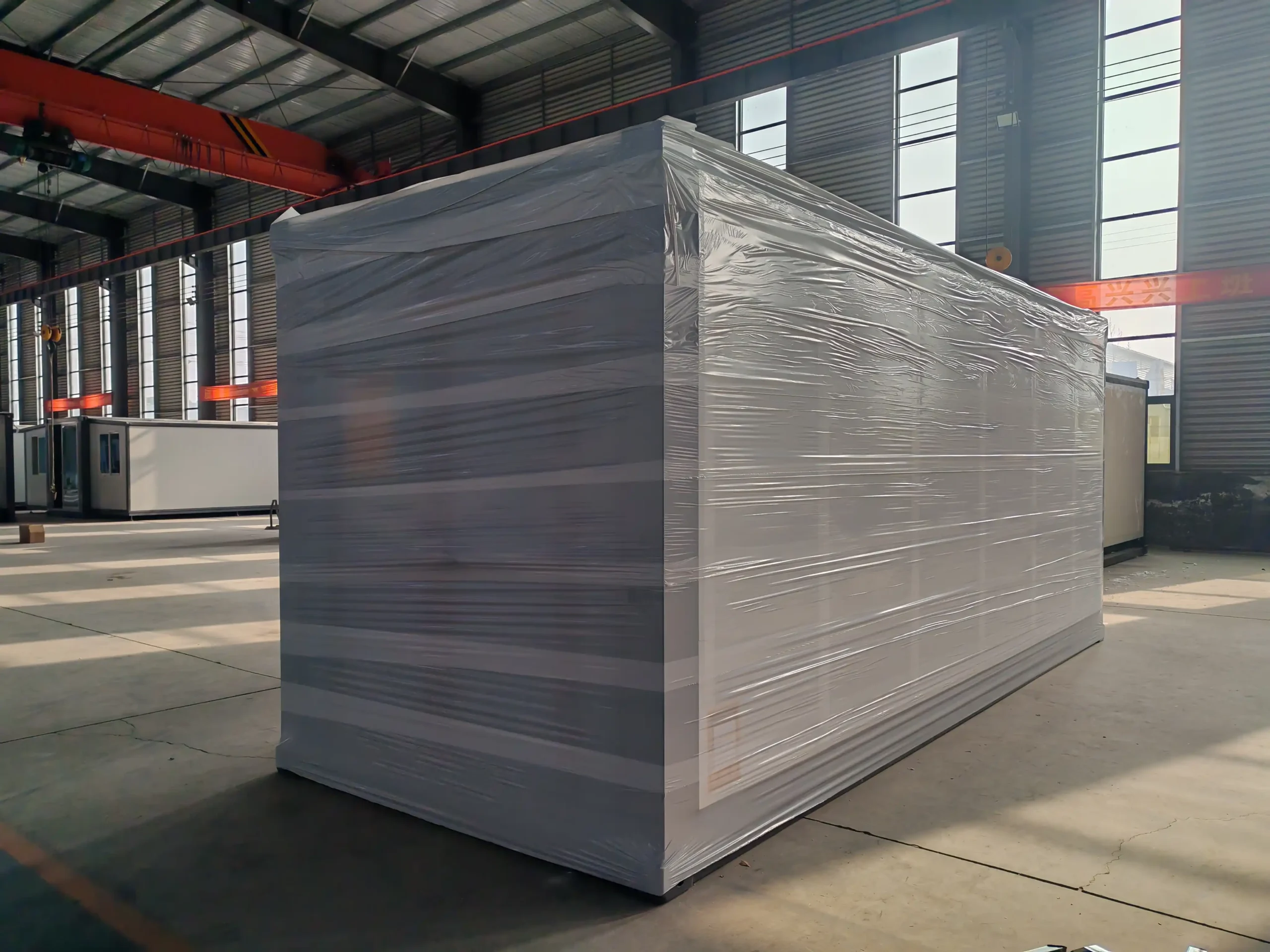 A factory-wrapped expandable container house unit sitting on a warehouse floor, protected by multiple layers of clear stretch film packaging, ready for B2B export shipment.