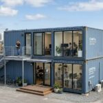 Aufbau: Zwei übereinander gestapelte blaue Container, ausgestattet mit bodentiefen Fenstern und Schiebetüren, die einen hellen, modernen Innenraum mit Schreibtischen und Personal freigeben. Zugänglichkeit: Eine äußere Metalltreppe führt zu einem Balkon im zweiten Stock, auf dem eine Person steht. Der Eingang im Erdgeschoss verfügt über eine kleine Holzterrasse und Kübelpflanzen. Umgebung: Gelegen auf einem Kiesplatz in der Nähe eines Hafens, mit Frachtschiffen und einer Stadtsilhouette im Hintergrund. Mehrere weiße Klimageräte sind am Sockel montiert, und Baufahrzeuge stehen in der Nähe. Kontext: Ein praktisches und ästhetisches Beispiel für umgenutzte Containerarchitektur, die als hochwertiges mobiles Baustellenbüro dient.
